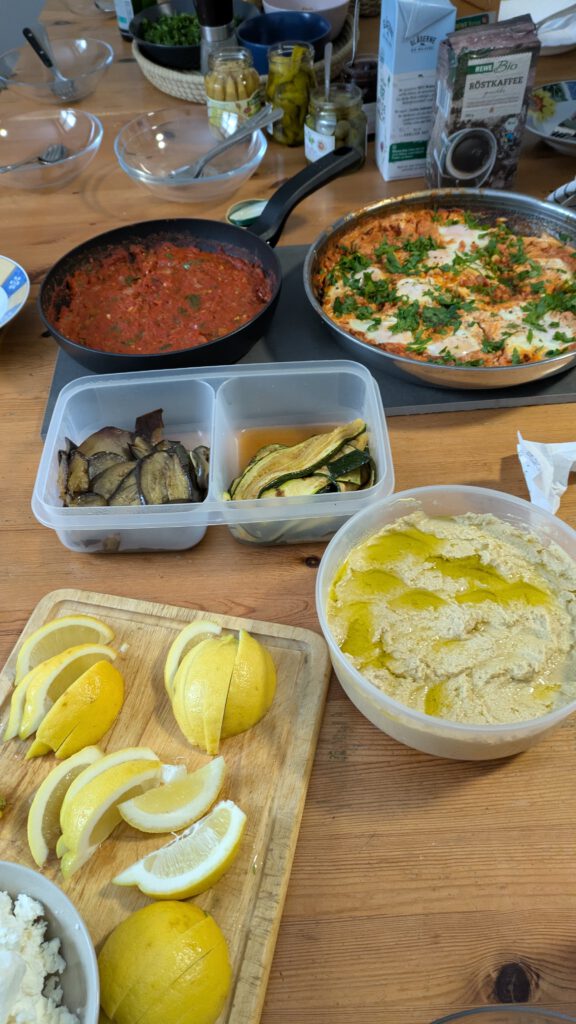 Ein gedeckter Tisch mit Humus, Zitronenspalten, Antipasti, WeiĂkĂ€se und Shakshuka. dazwischen Teller, Besteck und 2 Milchpackungen