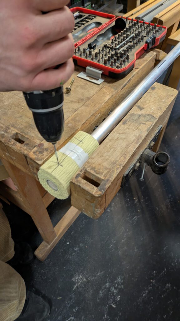 In einer Werkbank ist heller um einen Stab gewickelter Stoff eingespannt. Ein jemand bohrt ein Loch an eine von zwei markierten Stellen auf dem Stoff,