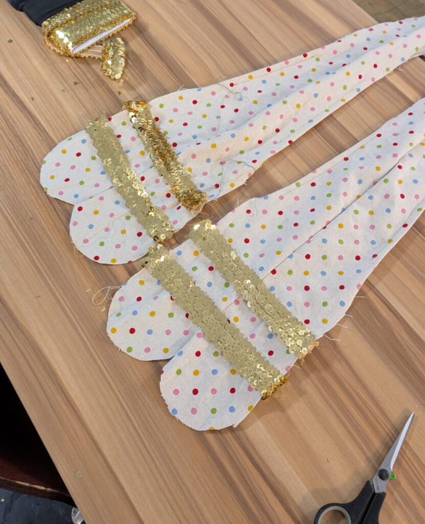 Auf einem Tisch liegen Zwei bunte gepunktete Stoffteile in der Form eines langen Tropfens. Quer aufgenÀht goldenen Pailletten Streifen