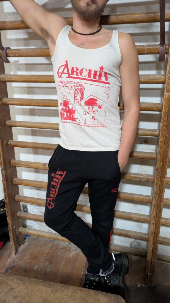 eine männliche Person steht vor einer Wand, sie trägt ein ärmelloses Top und eine Jogginhose mit roten Streifen auf denen Archiv gedruckt ist